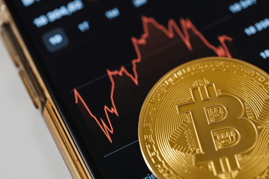 Ao fundo um smartphone com gráficos de criptomoedas. A frente uma moeda de Bitcoin (BTC) em dourado.