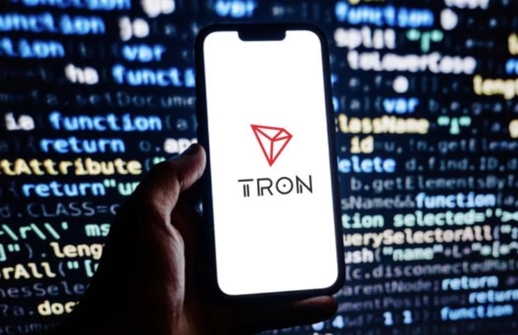 Fundo com dados em uma telão. Mão segurando smartphone. Na tela o símbolo da Tron (TRX).